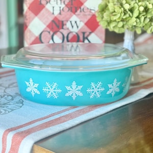 May include: A turquoise glass casserole dish with a clear glass lid. The exterior of the dish features a repeating white snowflake pattern around the sides. The dish has integrated handles on both sides and a smooth, rounded base design.