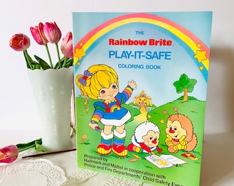Puede incluir: Un libro para colorear vintage de Rainbow Brite con un arcoíris, estrellas y los personajes Rainbow Brite, Twink y Grumpy. La portada del libro tiene el título "The Rainbow Brite. Play-It-Safe Coloring Book" y el texto "Prepared by Hallmark and Mattel in cooperation with Police and Fire Departments' Child Safety Experts".