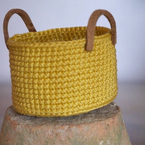 Puede incluir: Una pequeña cesta de ganchillo amarillo mostaza con dos asas de cuero marrón. La cesta es cilíndrica y se apoya sobre una superficie de terracota. El punto de ganchillo crea una apariencia texturizada.