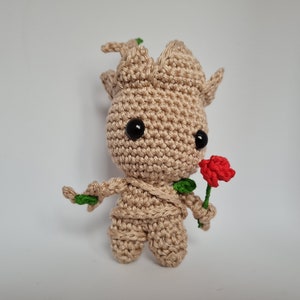 Puede incluir: Figura de ganchillo hecha a mano, de color beige, con grandes ojos negros y una rosa roja. La figura representa un personaje parecido a un árbol, sosteniendo la rosa. Tiene pequeñas hojas verdes.
