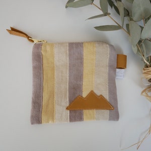 Op de afbeelding: Een gestreepte stoffen tas met een ritssluiting en een bruine leren berg-applicatie. De tas is gemaakt van lichtgrijs en geel gestreept stof.