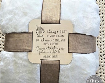 May include: A white faux fur blanket is tied with a brown ribbon and a wooden sign that reads "A house is built of walls & beams, but a home is made with hopes & dreams. Congratulations on your new place. Love, James & Kristi."