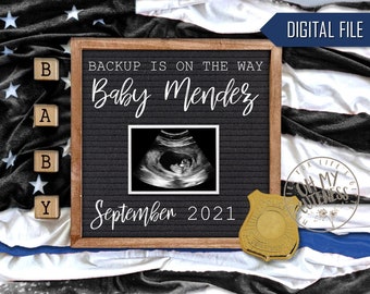 May include: A pregnancy announcement with a black letter board in a wooden frame. The board reads "Backup is on the way Baby Mendez September 2021" with an ultrasound image. The background features a blanket with stars and a police badge.