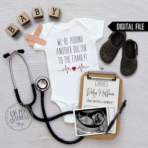 May include: A white onesie with the text "We're adding another doctor to the family!" and a red heart with a heartbeat line. A stethoscope, a clipboard with the text "2022 Baby Hoffman Due in December", a black and white ultrasound image, and two black clogs are also in the image.