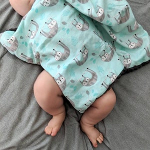 May include: A baby is swaddled in a soft, blue and gray blanket with a sloth pattern. The blanket is made of a lightweight fabric and has a soft, fuzzy trim.