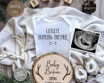 May include: A white onesie with the text "Littlest Hunting Partner" and two arrows. A wooden sign with the text "Baby Boone Coming October 202X" and antlers. A framed ultrasound image. A white blanket, antlers, and wooden blocks that spell "BABY".