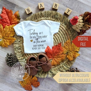 May include: A white babygrow with the text "Turkey ain't the only thing cookin' in the oven" with a gold heart and the text "Baby Hopkins Ready July 2021". The babygrow is surrounded by autumn leaves, a woven basket, and wooden blocks that spell out "Baby".