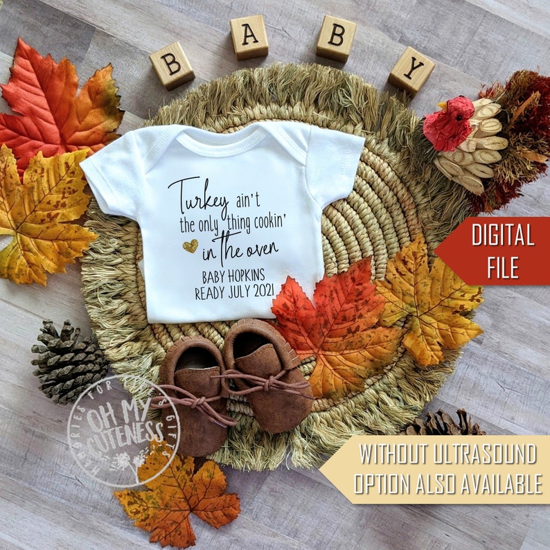 May include: A white babygrow with the text "Turkey ain't the only thing cookin' in the oven" with a gold heart and the text "Baby Hopkins Ready July 2021". The babygrow is surrounded by autumn leaves, a woven basket, and wooden blocks that spell out "Baby".