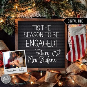 May include: A black letter board with white text that reads 'Tis the season to be engaged! Future Mrs. Bufana' with a ring emoji and a Polaroid photo of a couple holding hands. The letter board is surrounded by Christmas lights and a red and white stocking.