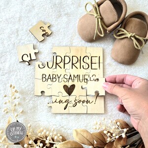 May include: Wooden puzzle pieces spelling out "Surprise! Baby Samuel is coming soon!" with a heart in the middle. The puzzle pieces are on a white fuzzy surface with two brown baby moccasins and a small sign that says "Oh My Cuteness".