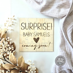 May include: A wooden puzzle piece announcement for a baby shower. The puzzle pieces are arranged to form the words "Surprise! Baby Samuels coming soon!" with a small heart in the middle.