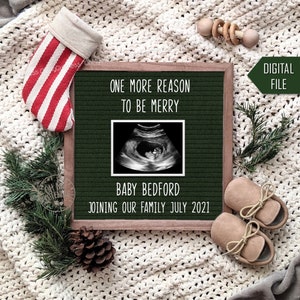 May include: A green letter board with the text "One more reason to be merry" and a sonogram image. Below the sonogram is the text "Baby Bedford joining our family July 2021". The letter board is surrounded by a white knitted blanket, a red and white striped stocking, a wooden toy, a pine branch, and a pine cone.