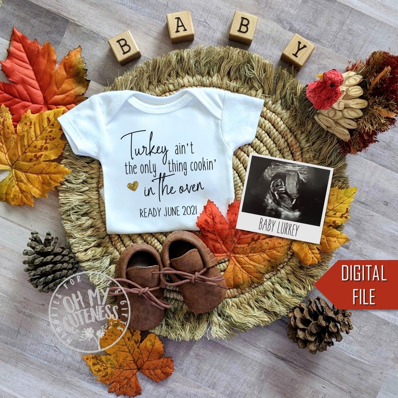 May include: A white babygrow with the text "Turkey ain't the only thing cookin' in the oven" and a heart with gold glitter. The babygrow is surrounded by autumn leaves, a small wooden turkey figurine, and a wooden block spelling "BABY".