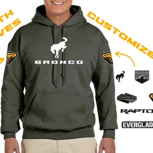 May include: A dark green hooded sweatshirt with a white Ford Bronco logo on the front. The sleeves have patches that can be customized with different designs, including "Wildtrak", "Raptor", "Everglades", "Black Diamond", "Outer Banks", and "Badlands".