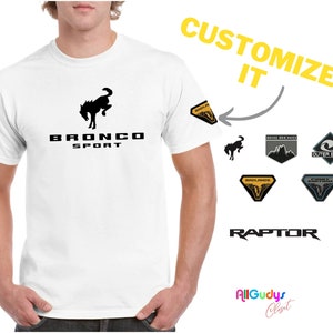 May include: White t-shirt with a black Ford Bronco Sport logo. The shirt has a customizable patch on the sleeve. The patch options include a black and white Bronco logo, a gray and black "Basecamp" logo, a gray and black "Outer Banks" logo, a yellow and black "Badlands" logo, and a yellow and black "Raptor" logo.
