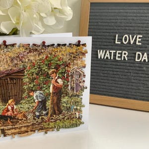 May include: A jigsaw puzzle depicting a garden scene with figures working amongst plants and a small shed. The puzzle is on a white card. A framed grey board displays the words "LOVE WATER DAILY".