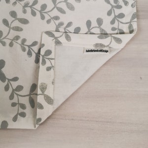 Può includere: Un runner da tavolo bianco con un motivo a foglie grigie. Il runner è piegato a metà e ha un'etichetta che dice "IsabellaHandcraftDesign".