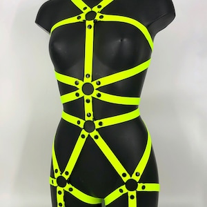 Può includere: Un body harness giallo neon con anelli e occhielli in metallo nero. Il harness ha cinghie che si incrociano sul petto, in vita e sulle gambe.