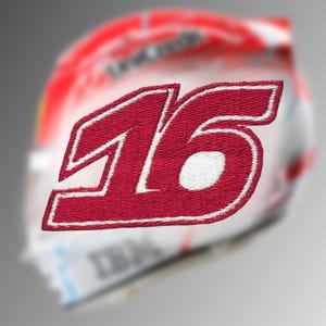 May include: A red embroidered patch featuring the number "16" in a bold, stylized font with a white border. The patch is likely for apparel or accessories, with a blurred background suggesting a helmet or similar item.