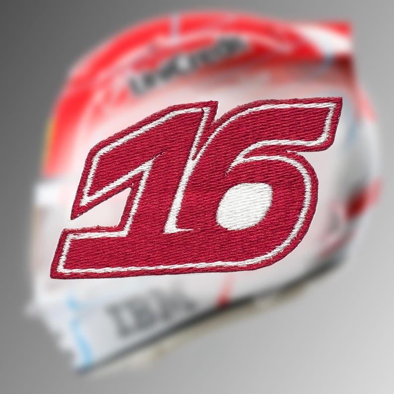 May include: A red embroidered patch featuring the number "16" in a bold, stylized font with a white border. The patch is likely for apparel or accessories, with a blurred background suggesting a helmet or similar item.