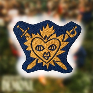 Könnte beinhalten: Ein gold gestickter Patch mit einer Herzform mit Augen und einem Mund, umgeben von einer Krone und zwei Schwertern. Der Patch befindet sich auf einem dunkelblauen Hintergrund.
