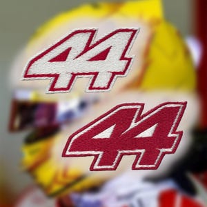 May include: Two embroidered patches featuring the number "44". The numbers are white with a red outline and are stitched onto a white background. The background is blurred, suggesting the patches are for a racing helmet or apparel.