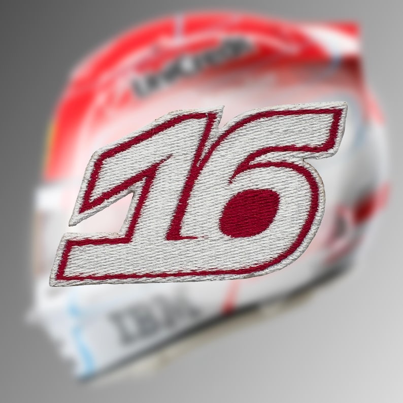 May include: Embroidered patch featuring the number "16" in white with a red outline and filling. The patch has a textured appearance, suggesting a fabric material. The background is blurred, showing a racing helmet.