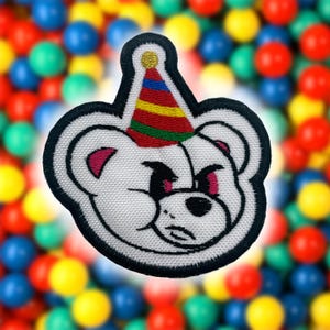 May include: White embroidered patch of a bear wearing a colorful party hat. The bear has black eyes and a black nose. The patch is surrounded by a black border.