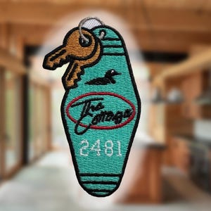 Op de afbeelding: Turquoise sleutelhanger met een gouden sleutel en een zwarte eendengrafiek. De tag leest "The Cottage" in rood script in een rode ovaal, en het nummer "2481" in wit. Een zilveren sleutelring is bevestigd.
