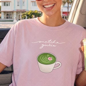 Puede incluir: Camiseta rosa claro con la frase "matcha girlie" y un dibujo de un matcha latte. La persona lleva gafas de sol rojas y sostiene una bebida verde en un vaso transparente con pajita.