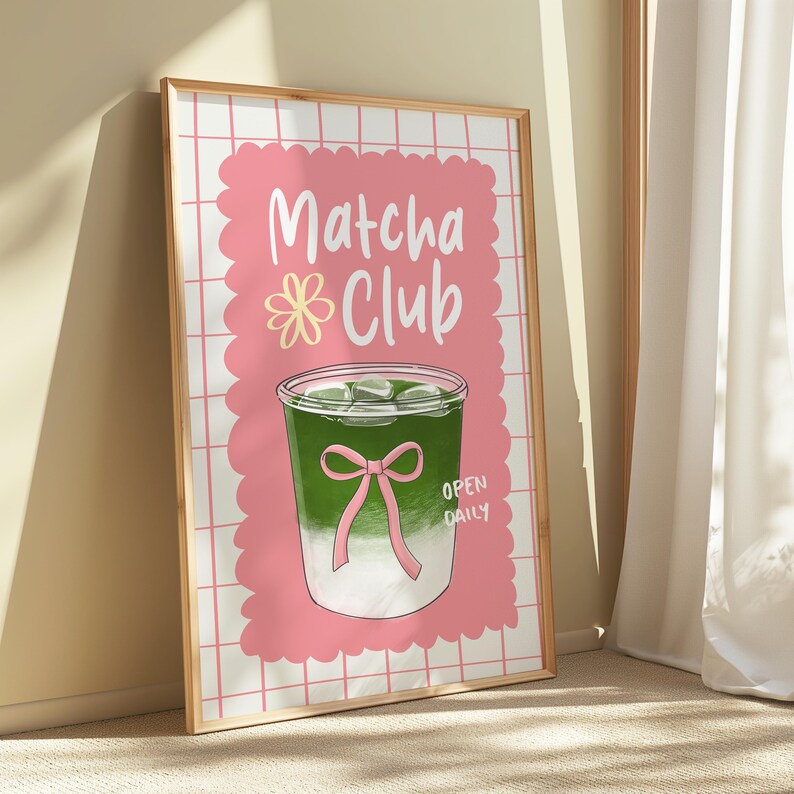 May include: A framed poster with a gold border features a pink background with the words "Matcha Club" in white script. A green matcha drink with ice and a pink bow is illustrated below the text. The words "Open Daily" are at the bottom.