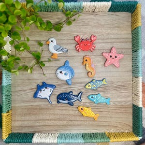 May include: A collection of ten small, flat, decorative sea-themed figures including a seagull, a red crab, a pink starfish, a seahorse, a stingray, a whale shark, and four various fish in blue, yellow, and patterned designs with cartoon faces.