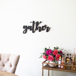 May include: Black wooden sign with the word "gather" in cursive script, hanging on a white wall. A gold bar cart with pink flowers and bottles is in the foreground.