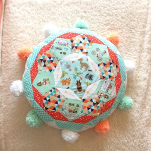 May include: A round, multi-colored patchwork cushion with a whimsical woodland animal design. The cushion is surrounded by white, peach, and light blue pom-poms.