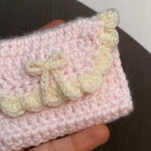 Puede incluir: Cartera de ganchillo hecha a mano en rosa suave con borde festoneado y lazo color crema. La cartera se sostiene en una mano, mostrando el delicado trabajo de puntadas y la combinación de colores. Un accesorio encantador.