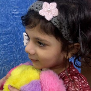 Puede incluir: Una diadema gris tejida a ganchillo con una flor rosa claro y un centro de perlas. La diadema la lleva una persona joven. La persona sostiene un animal de peluche colorido con pelaje amarillo, rosa y morado.