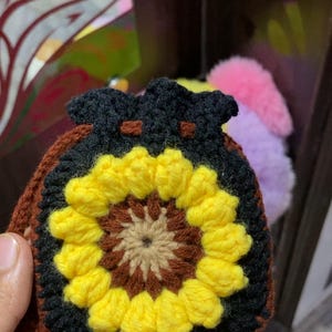 Puede incluir: Bolsa de ganchillo hecha a mano con un diseño de girasol. La bolsa tiene una base marrón, un borde negro y un girasol amarillo y marrón en el centro. Está hecha a mano y tiene un cierre de cordón.