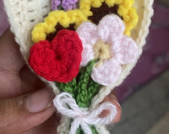 Puede incluir: Un ramo de ganchillo hecho a mano con un corazón rojo, girasoles amarillos, una flor rosa y una ramita morada. Las flores están envueltas en una envoltura de ganchillo color crema y atadas con un lazo blanco. Un pequeño anillo plateado está adjunto en la parte superior.
