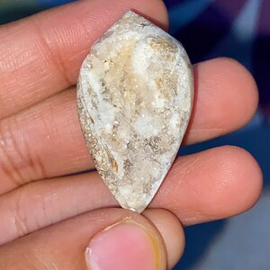 May include: A teardrop-shaped, natural stone with a rough, textured surface. The stone features a mix of beige, white, and light brown colors. The stone is held between fingers, showcasing its unique shape and natural patterns.