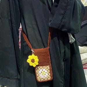 Puede incluir: Un bolso bandolera de ganchillo marrón para teléfono con un charm de girasol amarillo. El bolso presenta un diseño de granny square cuadrado multicolor en la parte delantera. La correa también está tejida.
