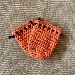 May include: Two crocheted orange pot holders with black trim. The pot holders are made with a simple stitch pattern and have a textured surface.