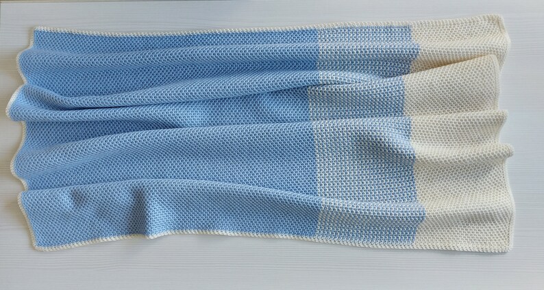 May include: A blue and white crocheted baby blanket with a textured pattern. The blanket is folded in half and has a white border.