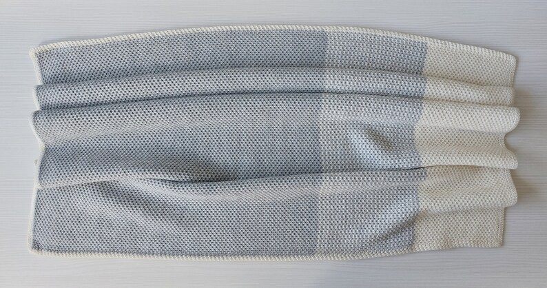 May include: A soft, gray and white knitted throw blanket with a textured pattern. The blanket is folded in half and has a white border.
