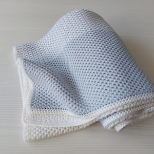 May include: A soft, gray and white knitted baby blanket. The blanket is folded and rolled up, showing the texture of the knit.