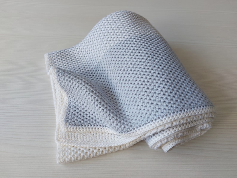 May include: A soft, gray and white knitted baby blanket. The blanket is folded and rolled up, showing the texture of the knit.