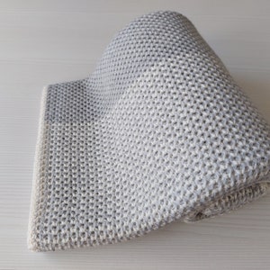 May include: A gray and white knitted throw blanket with a textured pattern. The blanket is folded in half and is lying on a white surface.