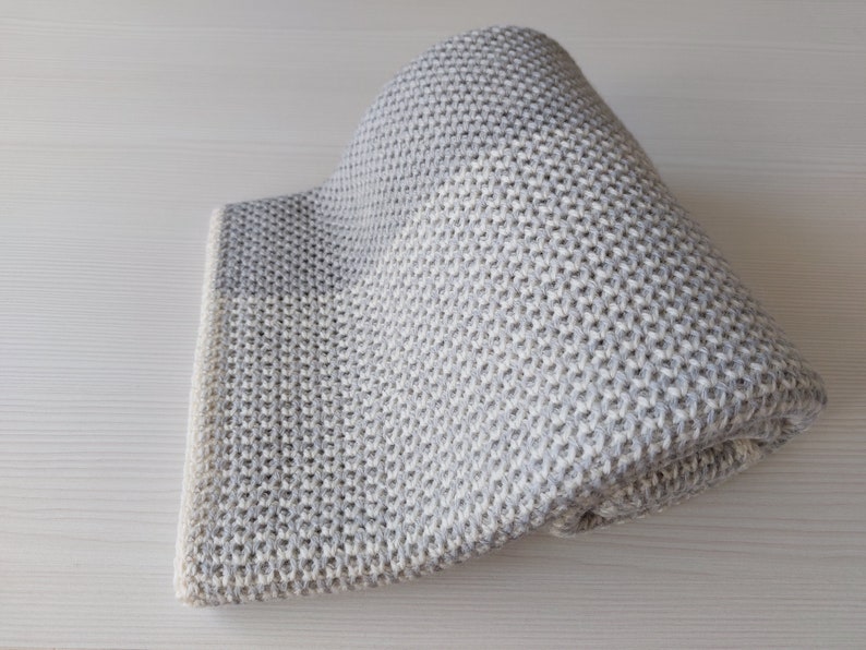 May include: A gray and white knitted throw blanket with a textured pattern. The blanket is folded in half and is lying on a white surface.