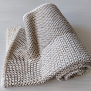 May include: A beige waffle weave throw blanket with a lighter beige stripe across the middle. The blanket is rolled up and is on a light wood surface.