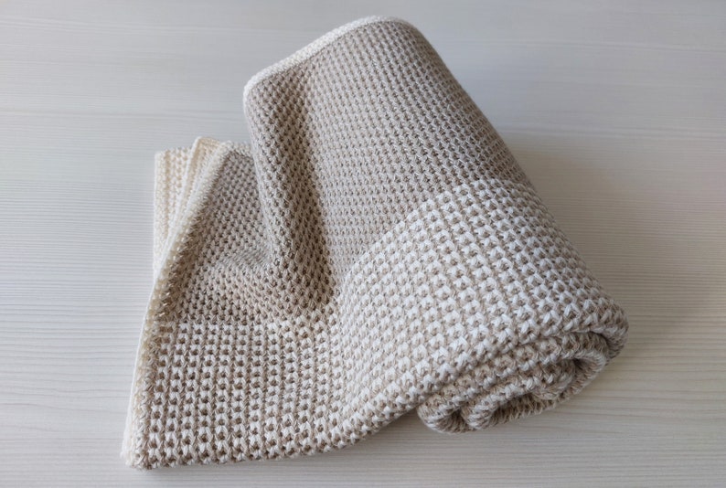 May include: A beige waffle weave throw blanket with a lighter beige stripe across the middle. The blanket is rolled up and is on a light wood surface.