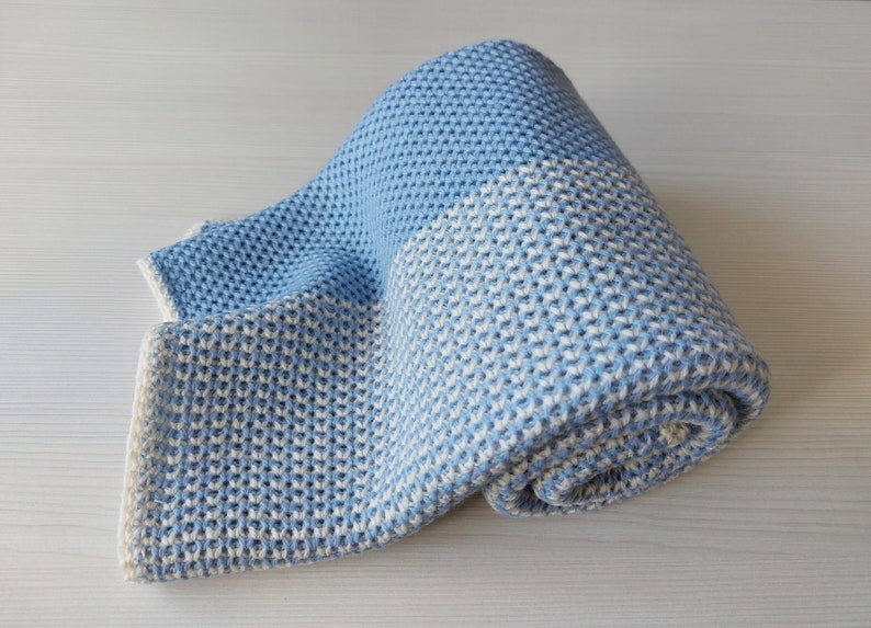 May include: A blue and white knitted baby blanket with a waffle-like texture. The blanket is rolled up and has a light blue color on one side and a white color on the other side.